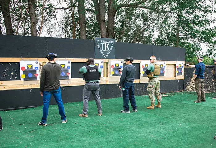 A group of men standing around looking at targets. One man is holding his gun up to the side of the wall and another man is pointing it towards the back wall.